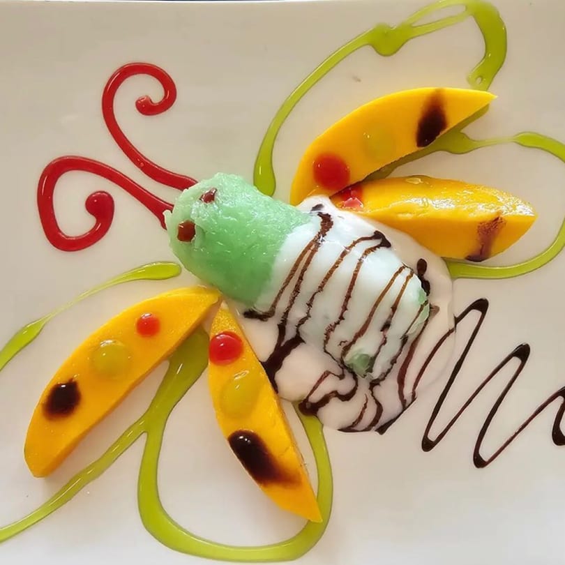 A butterfly-shaped dessert with green and white ice cream, mango slices, and colorful drizzles.
