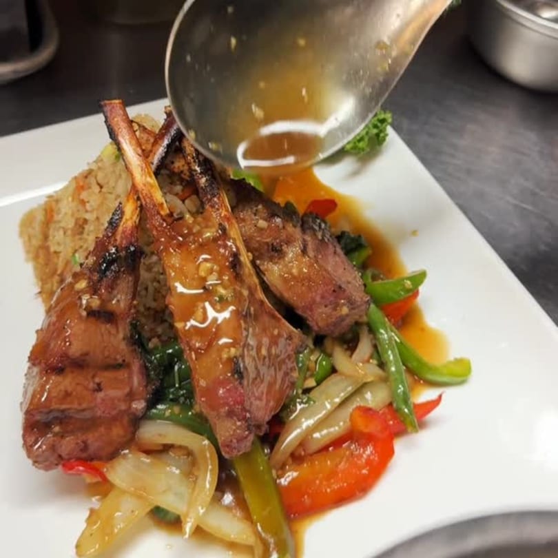 Grilled lamb chops with rice and stir-fried vegetables, being drizzled with sauce.